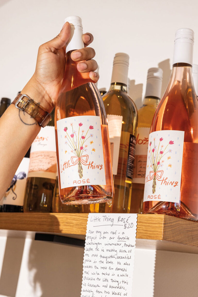 A hand holds a bottle of Little Thing Rosé wine on a wooden shelf, surrounded by other wine bottles. Below, a handwritten note describes the wine and its price of $20.
