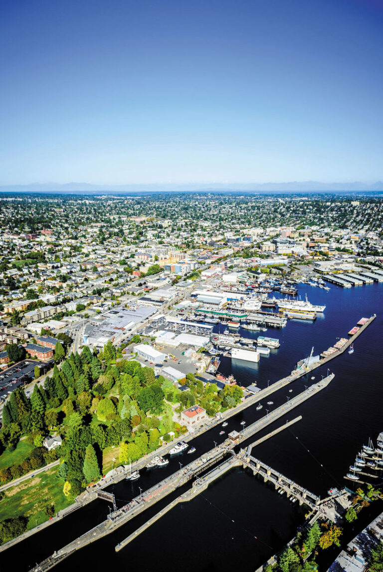 Aerial view of a city with a harbor, boats, green parks, and many buildings—featuring the vibrant Ballard neighborhood—all under a clear blue sky, stretching into the distance.