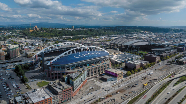 Aerial view of Lumen Field, a large stadium with a curved roof, nestled amid city buildings, roads, and greenery in Seattle’s SoDo neighborhood under a partly cloudy sky.