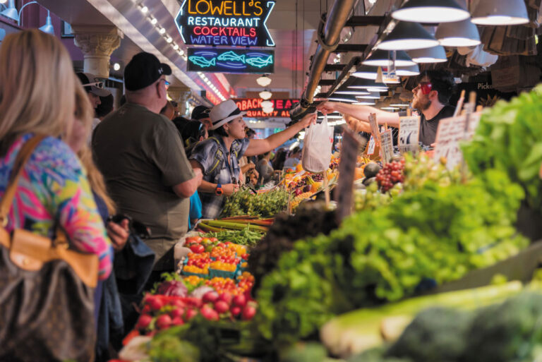 A busy downtown neighborhood Seattle market scene with people shopping for fresh vegetables and fruits. Colorful produce fills the foreground, while shoppers interact with vendors under bright lights and neon signs.