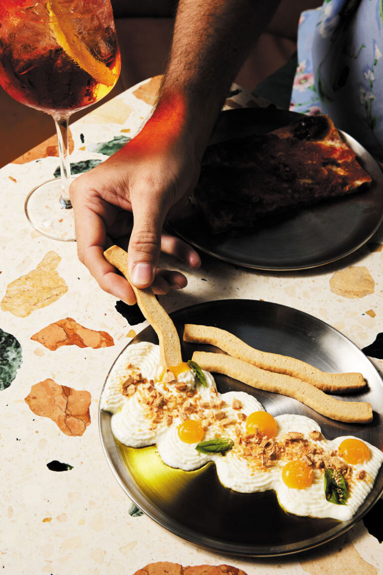 A hand dips a breadstick into a plate of whipped ricotta topped with herbs, nuts, and orange dollops. A glass of orange cocktail and a slice of pizza are on a terrazzo table.