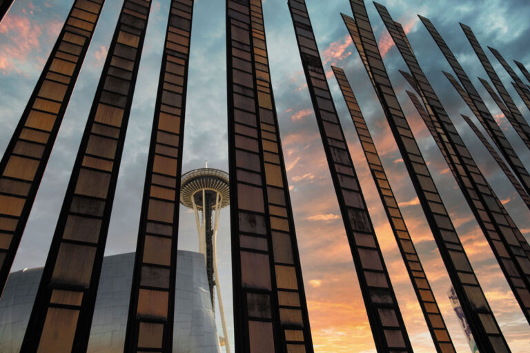 The Space Needle in Seattle is seen through vertical metal beams at sunset from the Queen Anne neighborhood, with a colorful sky and clouds in the background.
