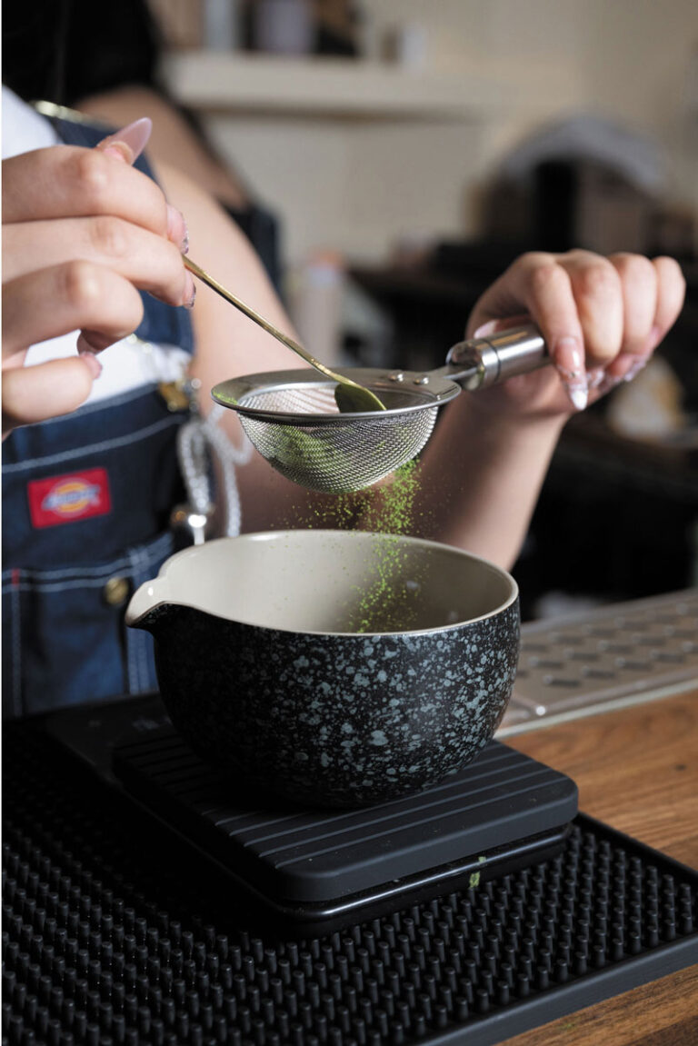 A person sifts green matcha powder through a small mesh strainer into a black speckled bowl on a wooden table, capturing the tranquil vibe of Seattle's Belltown neighborhood. Only their hands and part of their apron are visible.