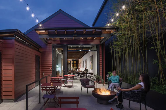A cozy outdoor patio at dusk with string lights, red chairs and tables, a fire pit, and two people talking. Nestled among bamboo plants in the SoDo Georgetown Seattle neighborhoods, the space leads into a warmly lit indoor area.
