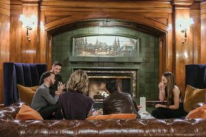 A group of people is seated on a brown leather couch in a cozy room with wood-paneled walls and a lit fireplace. They are engaged in conversation and enjoying drinks. Two wall sconces illuminate the scene, adding a warm ambiance.