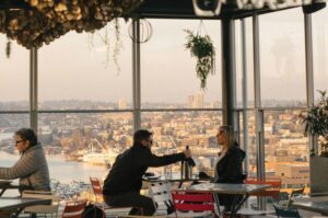 A cozy rooftop cafe with large windows overlooking a cityscape at sunset. Two people are seated at a table in front, engaged in conversation, and another person sits to the side. Hanging decorations and wine glasses add a warm ambiance.