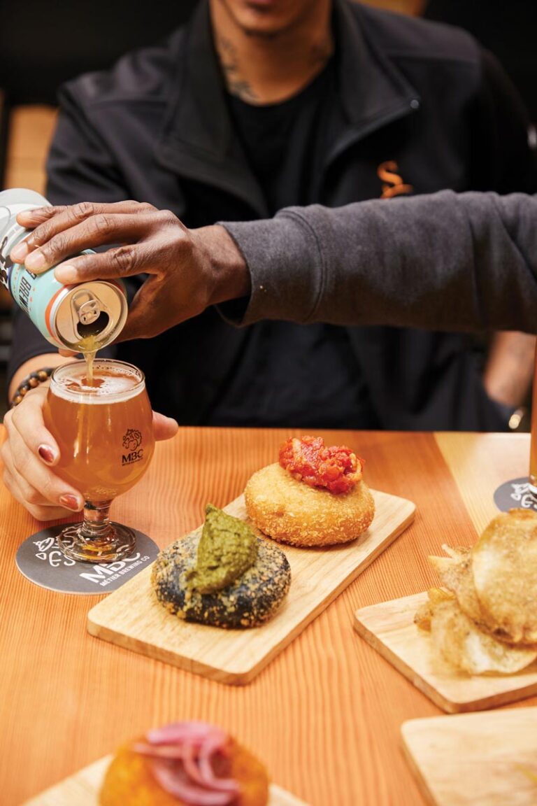 Two people sitting at a wooden table enjoy a drink being poured into a glass. In front of them are assorted breaded snacks with different toppings on wooden boards.