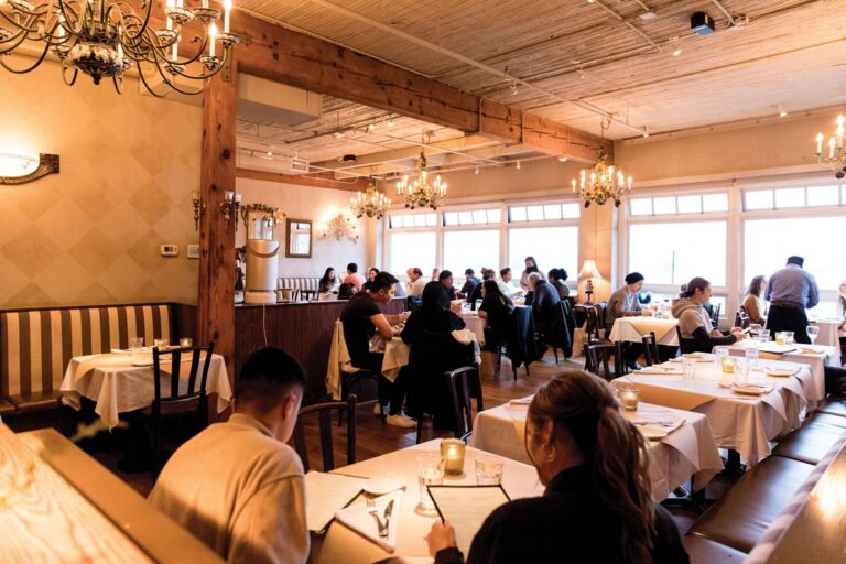 A cozy restaurant with wooden beams and chandeliers, filled with people dining at tables. The space is warmly lit, with large windows letting in natural light. The atmosphere appears lively and inviting.