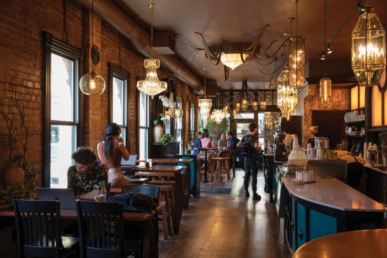 A cozy café with exposed brick walls and warm lighting. Patrons sit at tables and a person takes photos. Elegant chandeliers hang from the ceiling. The ambiance is welcoming and intimate, with people enjoying their time.