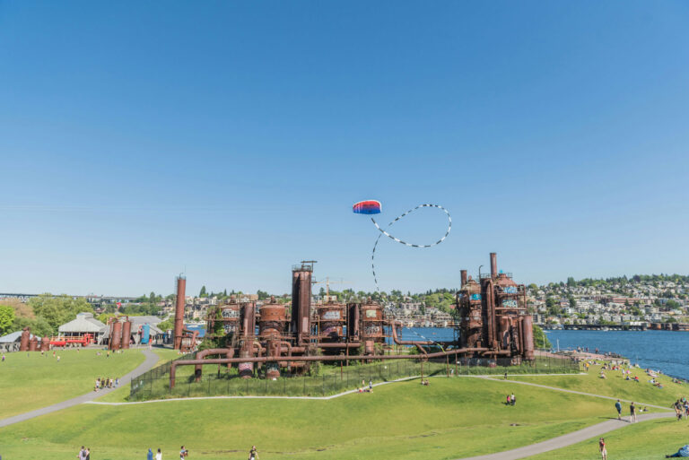 A sunny day at Gas Works Park with people relaxing on the green lawns. An industrial structure stands in the center, and a colorful kite flies high in the clear blue sky. A lake and cityscape are visible in the background.