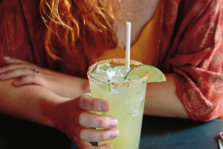 In the vibrant Georgetown neighborhood of Seattle, a red-haired person enjoys an iced drink in a glass with a salted rim and lime wedge. Nearby, a bowl of salsa and tortilla chips sits on the table. The person dons a red patterned top over a yellow shirt, capturing the area's lively essence.