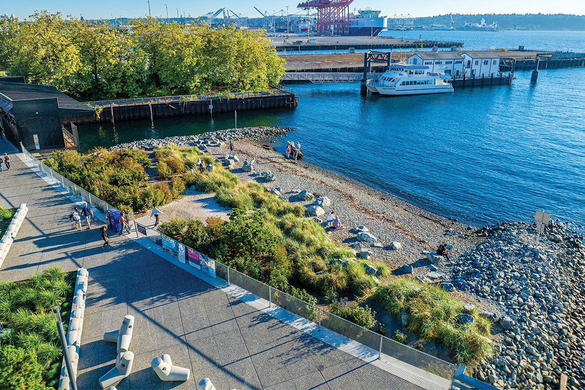 Aerial view of people walking along a landscaped waterfront park with greenery, a rocky shoreline, a docked white ferry boat, and an industrial port in the background under a clear blue sky.