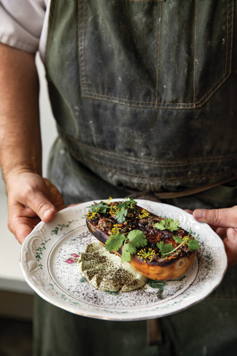 A person in an apron holds a decorated plate with roasted squash halves topped with greens and herbs. The dish is garnished with a creamy and herbaceous sauce on the side.