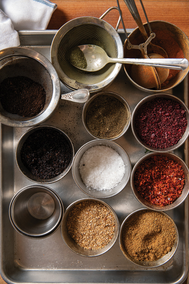 A tray filled with small bowls containing various spices and ingredients, including ground herbs, chili flakes, coarse salt, and dark powders. A stainless steel strainer and spoon rest nearby, with a white cloth in the corner.