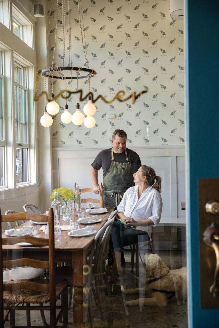A man in an apron stands smiling at a woman seated at a dining table set with plates and glasses. The restaurant interior has floral wallpaper, large windows, and a hanging light fixture. A dog lies on the floor next to the woman.
