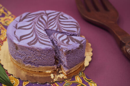 A round purple cake with a marbled pattern on top, with one slice slightly pulled out. It's placed on a golden base, with a decorative purple and yellow cloth beside it, and a large wooden serving utensil in the background.
