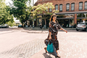 A woman wearing a black, red, and white dress and holding a blue bag walks across a brick street.