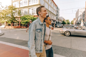 One person in a jean jacket with their arm around a person in a white shirt cross a colorfully painted street.