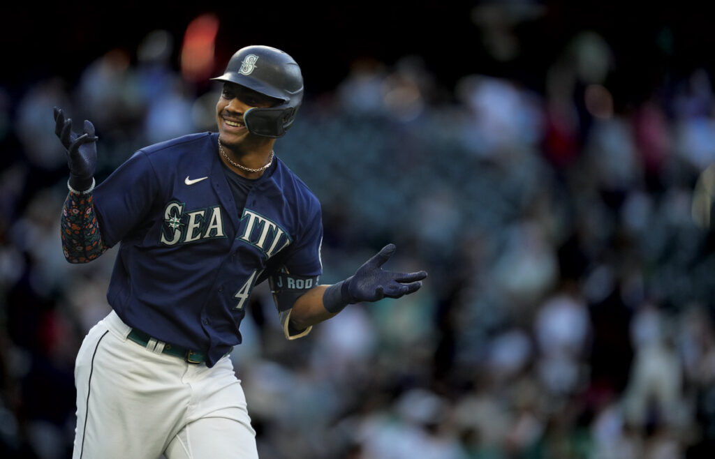 A photo of baseball player Julio Rodriguez in his Seattle Mariners uniform. He is wearing a dark blu shirt with the Nike logo and word Seattle across the chest. He has on white pants, navy blue gloves, and a navy blue hat. The background is a blurred out crowd.