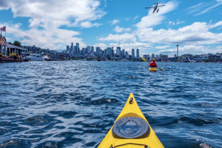 A view from a yellow kayak on a lake, approaching the Seattle skyline. A person is kayaking ahead, and a seaplane is flying above. The Space Needle is visible on the right, with boats docked along the shorelines.