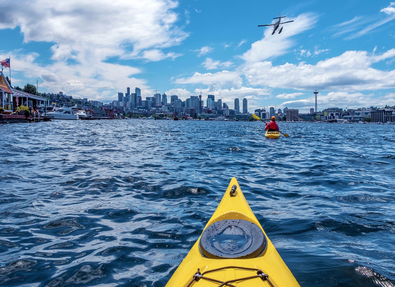 A view from a yellow kayak on a lake, approaching the Seattle skyline. A person is kayaking ahead, and a seaplane is flying above. The Space Needle is visible on the right, with boats docked along the shorelines.