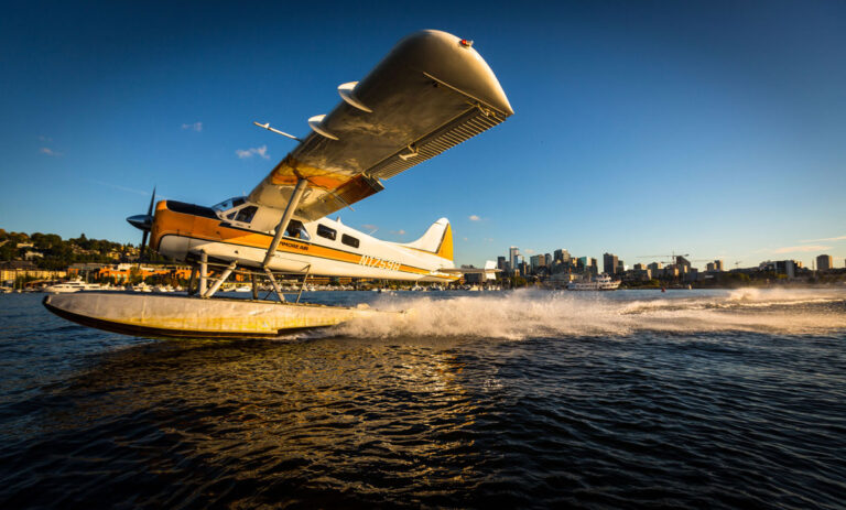 A seaplane takes off from the shimmering waters, creating a splash in its wake—a quintessential experience among things to do in Seattle. The city skyline stands tall in the background under a clear blue sky, bathed in warm sunlight.