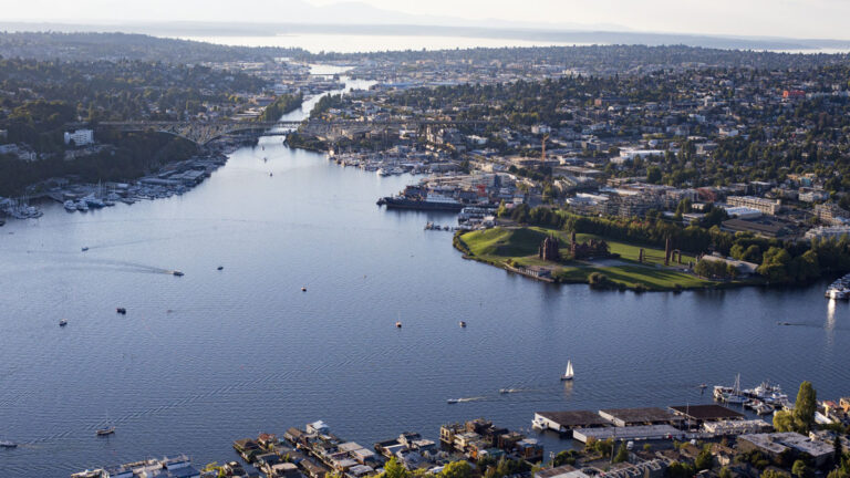 Aerial view of Seattle's cityscape with a large body of water flowing through. Boats dot the water, while a vibrant green area with buildings lines the shoreline. The city extends into the distance, offering an inviting blend of urban exploration and natural beauty—perfect for discovering things to do in Seattle.