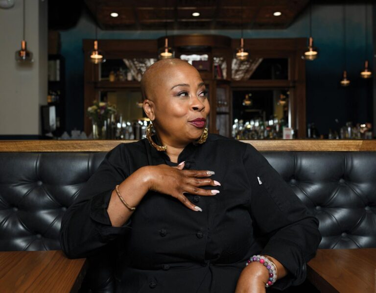 A chef in a black uniform sits in a restaurant booth with tufted leather seats. She has a bald head and wears gold earrings and bracelets. She holds her hand on her chest and gazes to the side, with a warm, ambient bar setting in the background.