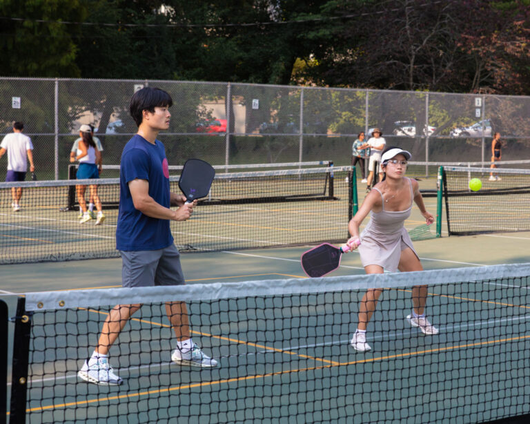 Two people play doubles pickleball on an outdoor court, both holding paddles and focusing on the game. Other players are active on nearby courts, and trees are visible in the background.