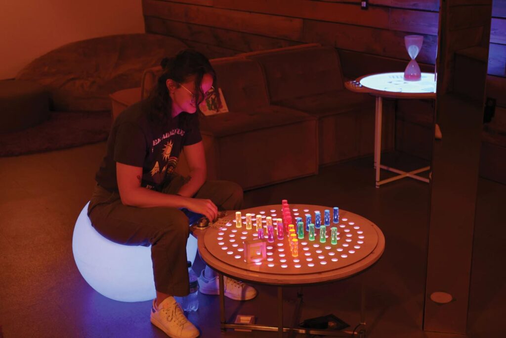 A person sitting on a glowing orb plays a colorful, illuminated board game on a round table in a dimly lit room with a couch and hourglass in the background.