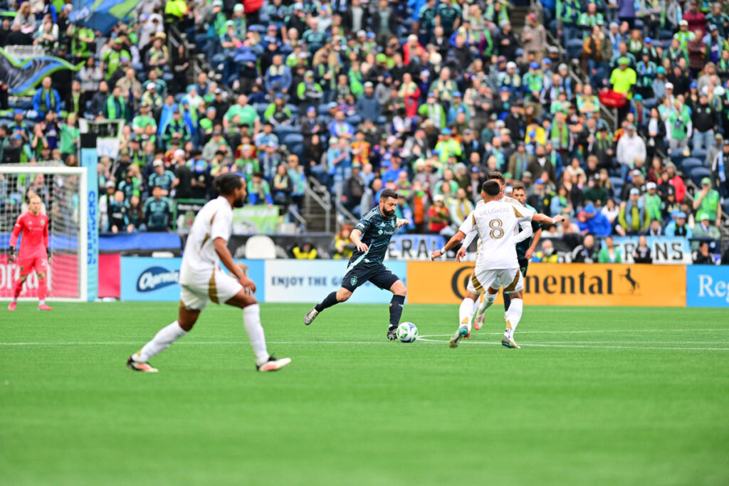 A soccer player in a blue uniform dribbles the ball past opponents in white during a match, with a crowd of fans filling the stadium seats in the background.