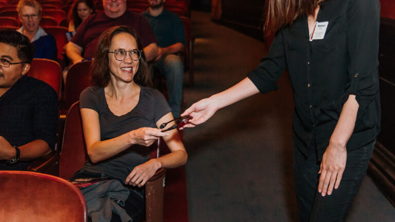 A woman sitting in a theater seat smiles while another person hands her a pair of 3D glasses. Other people are seated around them, and the environment appears friendly and casual.