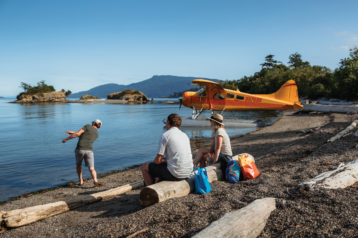 Three people sit on a a sandy beach with blue water, islands, and a yellow sealpane in the background.