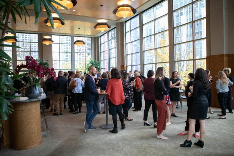 A group of people mingles at a networking event in a spacious room with large windows. Potted plants and hanging lights decorate the area. Most attendees are engaged in conversation, holding drinks.