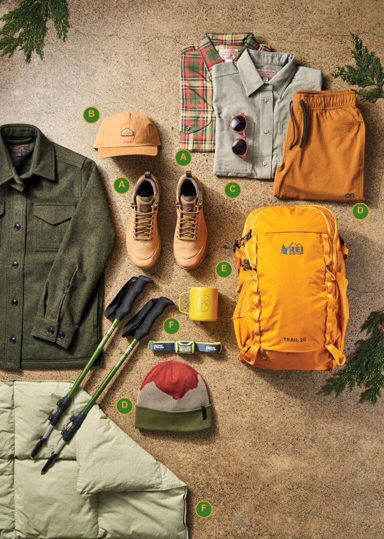 Camping gear flat lay: green wool jacket, tan hiking boots, plaid and gray shirts, tan cap, sunglasses, orange shorts, trekking poles, yellow mug, orange backpack, beanie, green quilted sleeping bag on a stone background with pine branches.