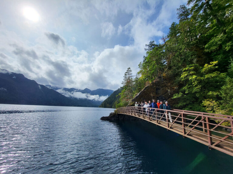 A group of people stroll along a wooden bridge over a serene lake, surrounded by lush green trees and mountains—a tranquil escape that feels like one of the peaceful things to do in Seattle. The sky is partly cloudy, with sunlight peeking through.