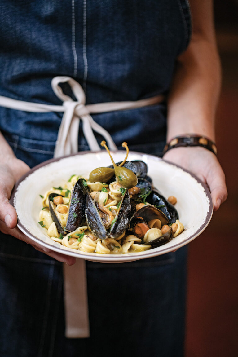A person in a blue apron holds a white plate filled with pasta, mussels, and garnished with caperberries and fresh herbs.