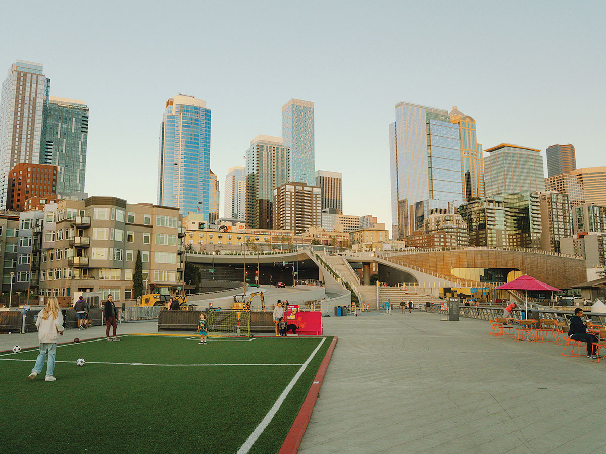 People play soccer and walk on a rooftop field in an urban park, with modern high-rise buildings and skyscrapers in the background under a clear sky. There are also tables and colorful seating on the right side.