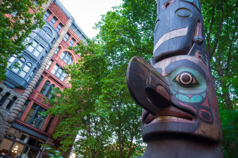 A close-up of a totem pole in front of a historic red and gray brick building, framed by green trees, highlights one of the captivating things to do in Seattle. The colorful, carved figures on the pole complement the building's windows and ornate details visible in the background.
