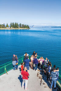 A group of people stand at the front of a ferry boat, looking out over blue water toward a distant tree-lined shoreline and a city skyline under a clear, sunny sky.