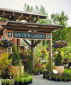 Outdoor entrance to Bayview Garden, with hanging baskets of colorful flowers, potted plants lining the walkway, and lush greenery in the background. The sign is mounted on a wooden structure.