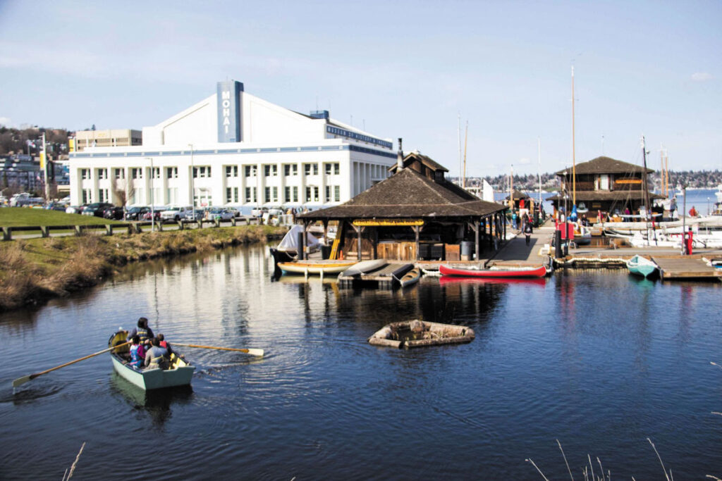 People row a small boat in a calm body of water near docks with wooden boathouses, canoes, and kayaks. A large white building with "MOHAI" signage is visible in the background. The day is clear and sunny.