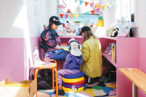 Two adults and a child sit together in a colorful, sunlit children’s reading nook with books, playful decorations, and bright wall colors. The child sits on stackable cushions while the adults read or talk nearby.