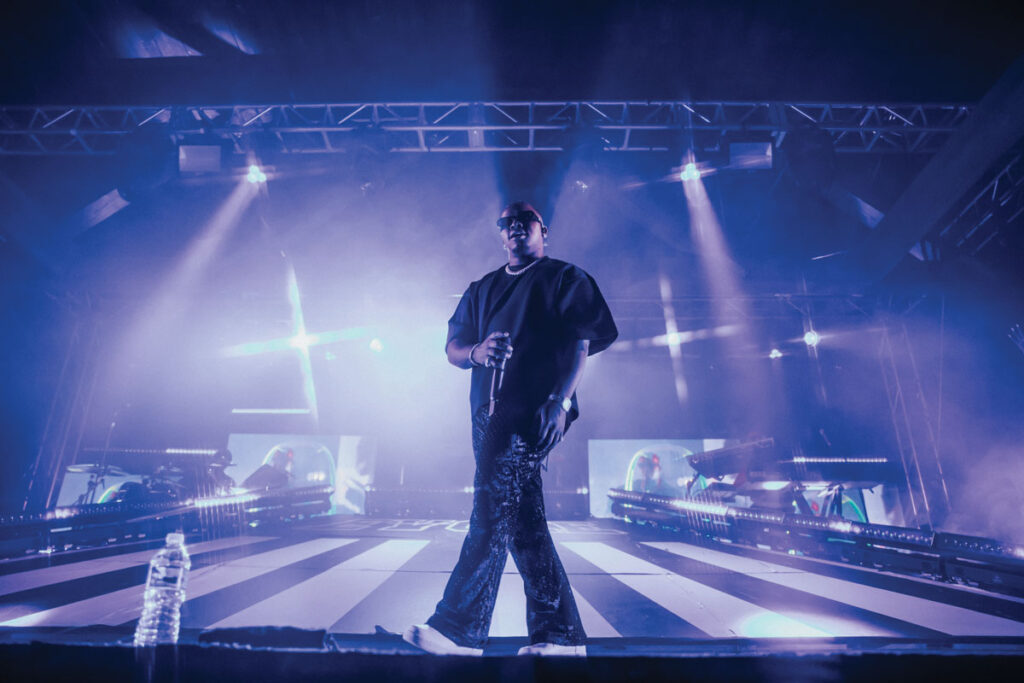A man wearing all black standings on a stage backlit by purple stage lights.