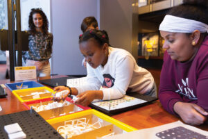 A young girl assembles pieces at a table with colorful trays of parts, while an adult woman and two other people watch in a bright, interactive learning space.