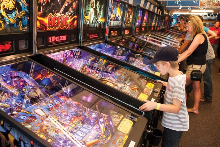 A young boy wearing a striped shirt and baseball cap plays a pinball machine in an arcade. Other people are also playing pinball machines lined up in the background. The arcade is bustling with activity.