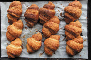 Ten golden-brown croissants on a baking sheet lined with parchment paper, fresh from the oven. The pastries have crisp, flaky layers and varying shapes and sizes.