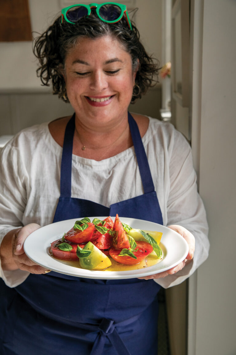A smiling person wearing a white shirt and blue apron holds a plate with a colorful tomato and basil salad.