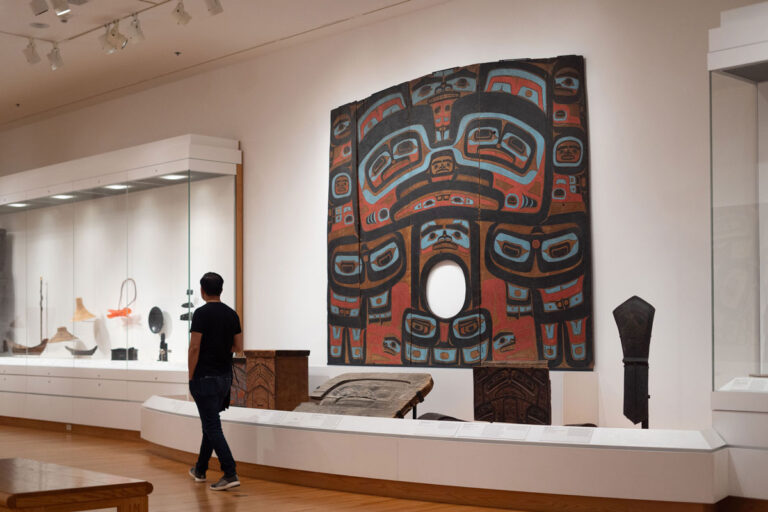 A person walks through a museum exhibit, observing a large, colorful indigenous tapestry with a circular opening in the center. Display cases with various artifacts are visible in the background, offering a glimpse of things to do in Seattle beyond its bustling cityscape.