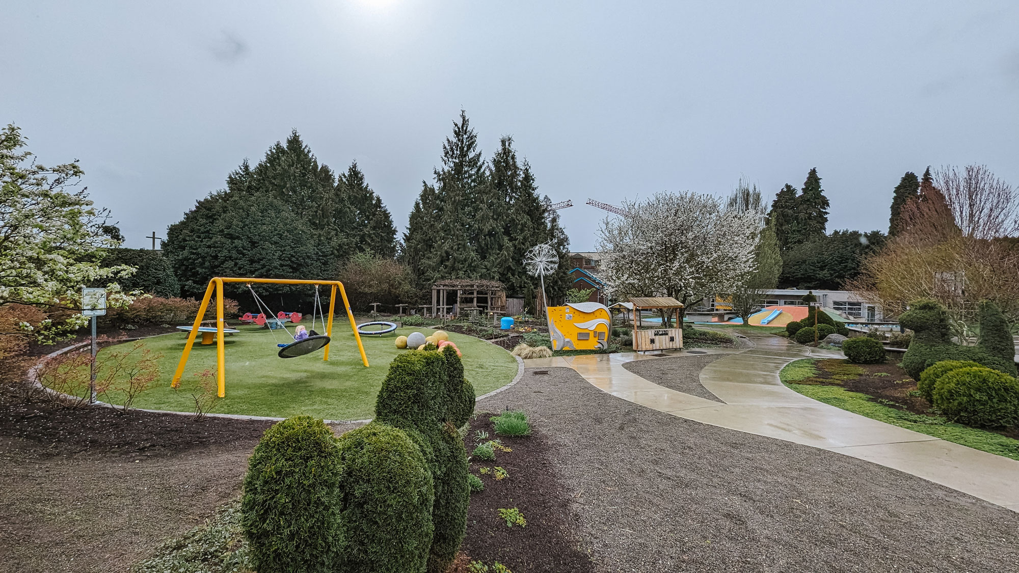 A yellow metal swing structure with a circular swing sits atop a green patch of grass that is shaped in a large circle. Other play structures are within the circle. To the right of the circular patch is a community pea patch, a yellow shed with organic metal details on the side, and a wooden stand. Trees border the park and a cement path winds through the area.